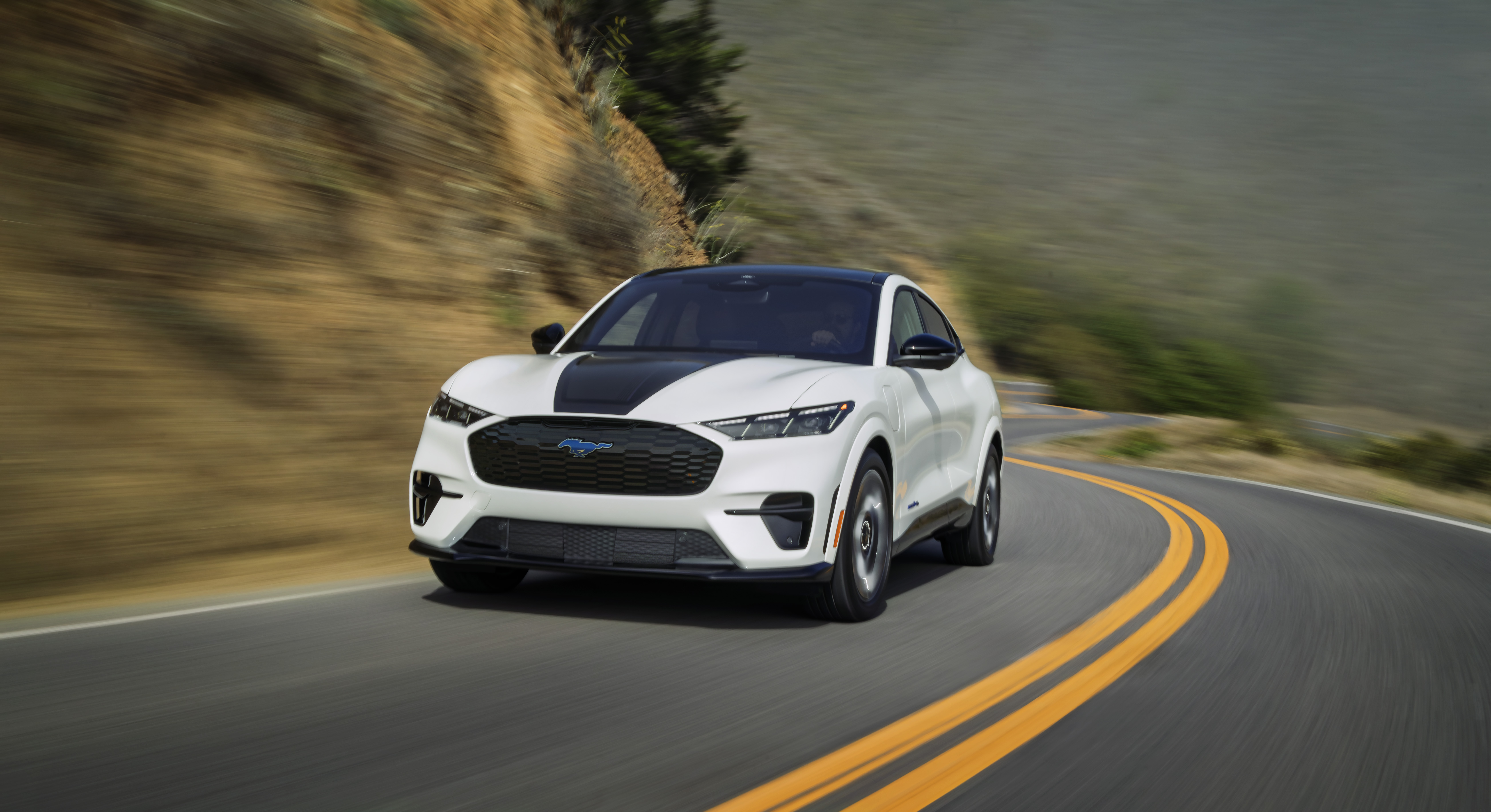 A white Mustang Mach-E drives up a hill. The background of a steep hill covered in trees blurs past.